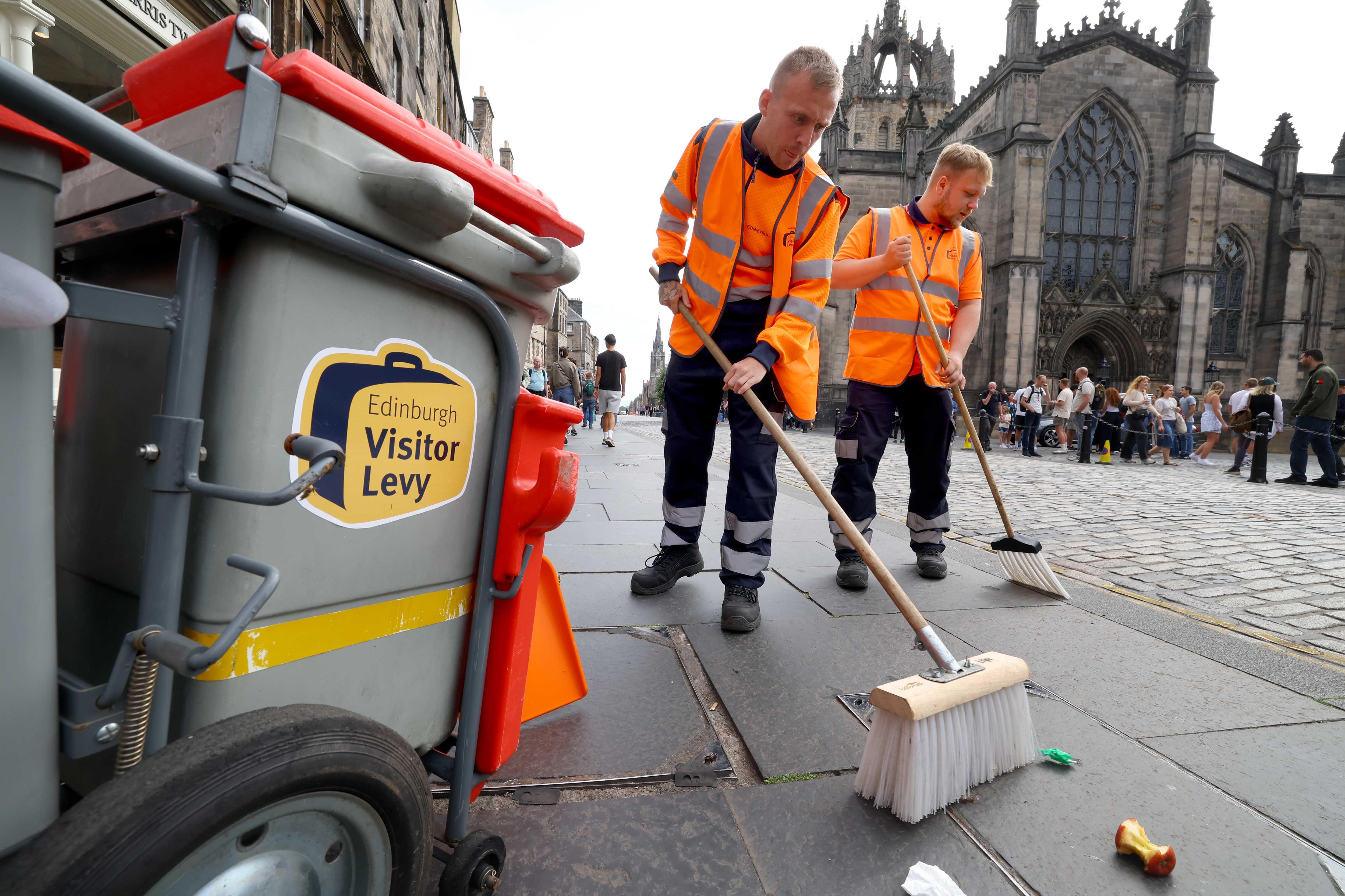 Two men in high-vis jackets sweeping the streets of Edinburgh next to a Visitor Levy logo on a bin