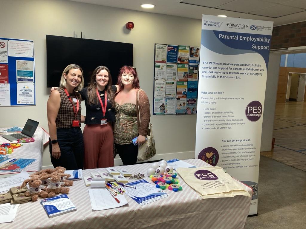Three women smiling in front of a Parental Employability Support stand.